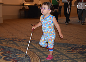A blind toddler smiles as she skips along with her long white cane.